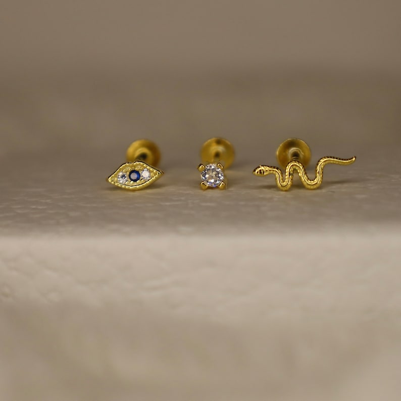 May include: Three gold body jewelry pieces with different designs. The first piece is a small eye with blue and white stones. The second piece is a small round stone. The third piece is a small snake.