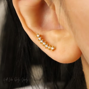 May include: A gold ear cuff with a row of small, clear cubic zirconia stones. The ear cuff is worn on the upper lobe of the ear.