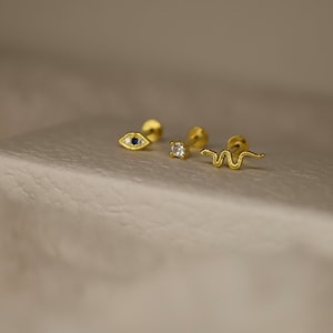 May include: Three gold body jewelry pieces with different designs. The first piece is a small eye with a blue stone. The second piece is a small round stone. The third piece is a small snake.