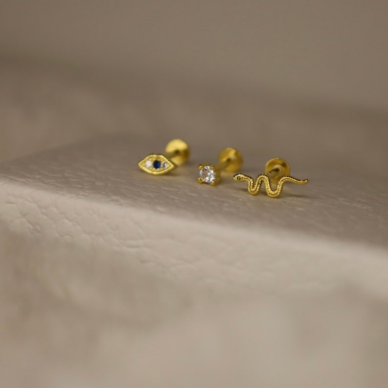 May include: Three gold body jewelry pieces with different designs. The first piece is a small eye with a blue stone. The second piece is a small round stone. The third piece is a small snake.
