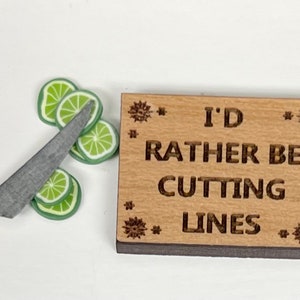 May include: A wooden cutting board with the text "I'd rather be cutting lines" engraved on it. A yellow handled knife and lime slices are on the surface next to the board.