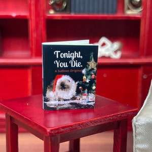 May include: Miniature book titled "Tonight, You Die" with a cat wearing a Santa hat on the cover. The book is on a small red table, with a red bookshelf in the background. A Christmas tree is also on the cover.
