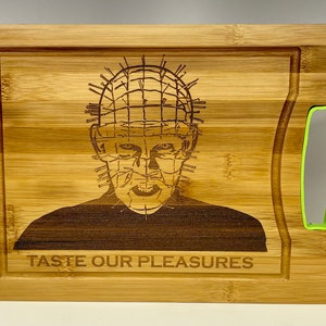 May include: A wooden cutting board with a green handle. The board has a laser-engraved image of a character from the movie Hellraiser, with the text "Taste Our Pleasures".