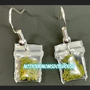 May include: A pair of novelty earrings featuring small, clear plastic bags filled with green material. The earrings have silver-colored hooks and the text "NOTYOURMOMSDOLLHOUSE" is visible on the bags.