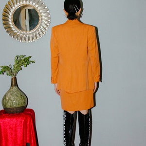 May include: A woman wearing a bright orange skirt suit with a matching blazer and skirt. She is wearing black knee-high boots with the text "FOR WALKING" on the side.