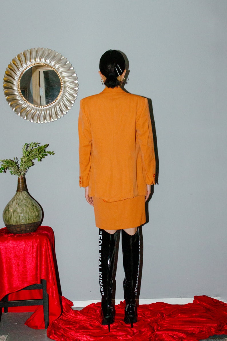 May include: A woman wearing a bright orange skirt suit with a matching blazer and skirt. She is wearing black knee-high boots with the text "FOR WALKING" on the side.