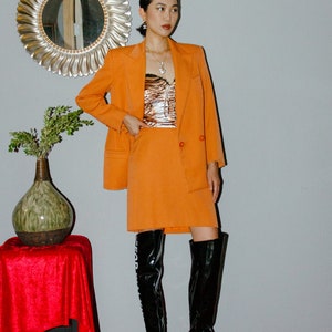 May include: A woman wearing a bright orange blazer and skirt suit with a black and white animal print top. She is wearing black thigh-high boots with a white graphic on the side.