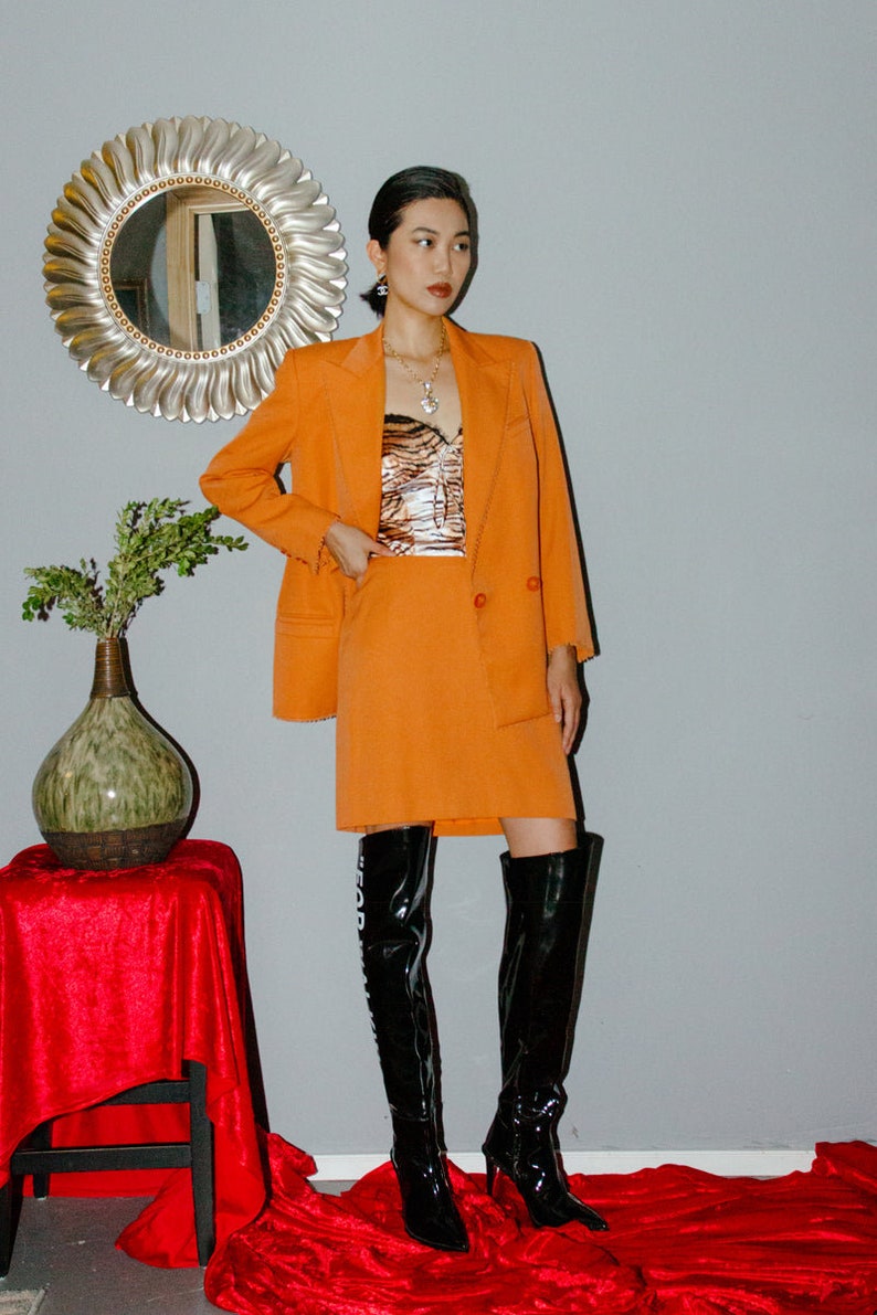 May include: A woman wearing a bright orange blazer and skirt suit with a black and white animal print top. She is wearing black thigh-high boots with a white graphic on the side.