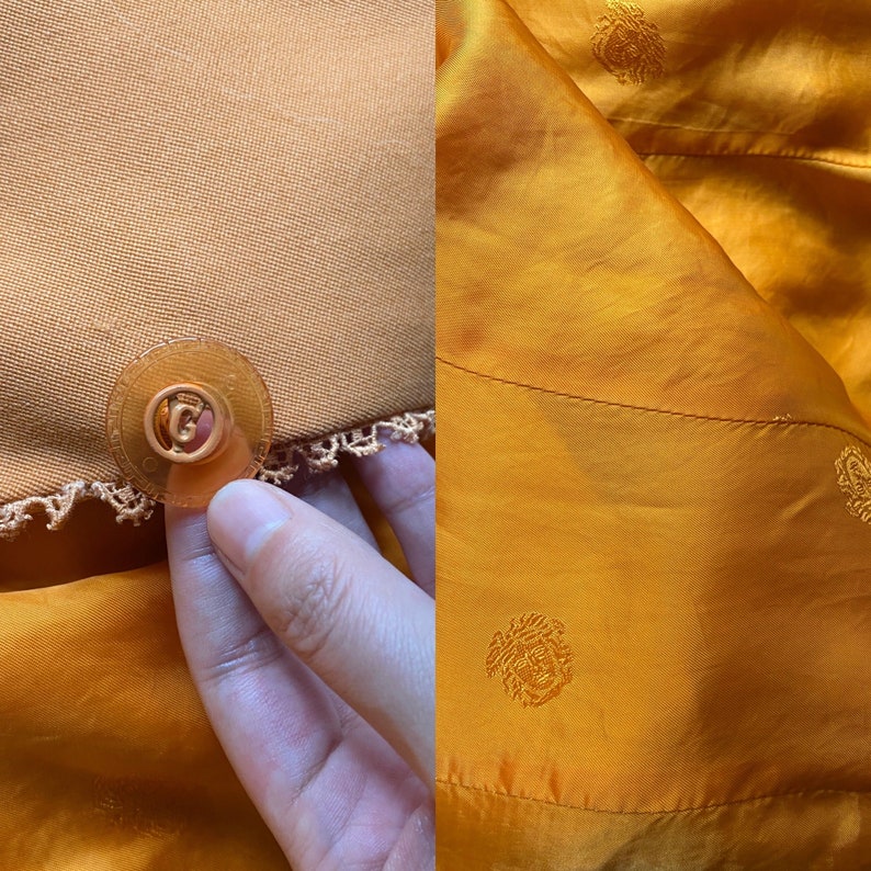 May include: Close-up of a gold button with a decorative edge, attached to a piece of orange fabric with a crocheted trim. The fabric is folded over, revealing a similar fabric with a subtle embossed pattern.