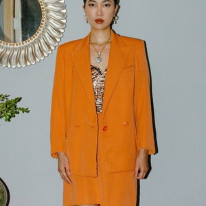 May include: A woman wearing a bright orange blazer and skirt suit with a scalloped trim. She is wearing a black and white animal print top underneath and a gold necklace with a heart pendant. She is also wearing black boots.