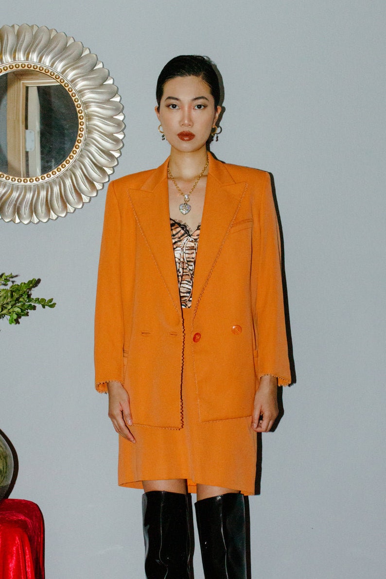May include: A woman wearing a bright orange blazer and skirt suit with a scalloped trim. She is wearing a black and white animal print top underneath and a gold necklace with a heart pendant. She is also wearing black boots.