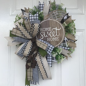 May include: A round decorative wreath featuring a central circular wooden sign with the text HOME sweet HOME in white script. The wreath is composed of layered mesh, green foliage, and multiple ribbons in black and white plaid, tan stripes, and solid brown.
