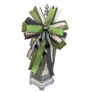 May include: A decorative lantern topper featuring a large bow with green, black, and tan ribbons. The bow has a variety of patterns, including polka dots and stripes. The lantern is silver and has a classic design.