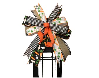 May include: A decorative bow featuring a mix of ribbons in various patterns and colors. The bow includes black and white gingham, orange, and ribbons with carrot and clover designs. The bow is likely for Easter or spring decor.