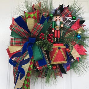 May include: A Christmas wreath featuring a nutcracker figure standing on a red drum. The wreath is decorated with green pine needles, a large multi-layered bow in red, green, blue, and gold plaid, and small red berry clusters and shiny ornaments.