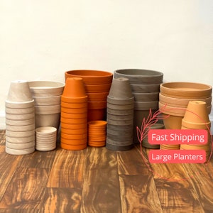 May include: A selection of terracotta plant pots in various sizes and colours, stacked and arranged on a wooden floor. The pots are brown, orange, and grey. The text "Fast Shipping" and "Large Planters" is visible in the image.