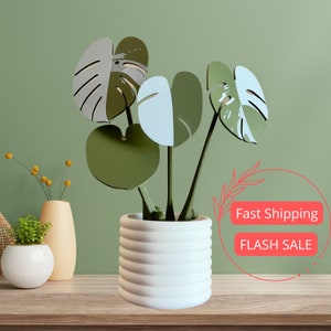 May include: A white ceramic planter with a ribbed design, holding a faux plant with green and white leaves. The plant is shaped like a monstera plant.