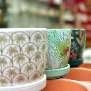 May include: Three ceramic plant pots with different tropical patterns. The pot on the left has a white background with gold palm trees, the middle pot has a light green background with white palm leaves, and the pot on the right has a dark green background with white leaves.