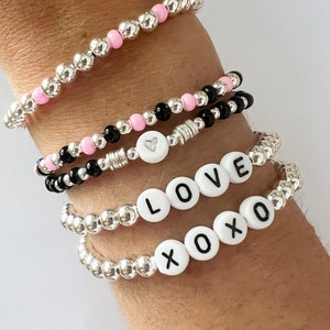 May include: A set of four silver bead bracelets with pink and black beads. One bracelet has a heart charm, and two bracelets have white letter beads that spell out "LOVE" and "XOXO".
