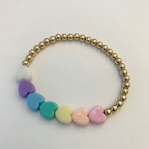 Op de afbeelding: Een gouden kralenarmband met pastelkleurige hartbedels. De bedels zijn in regenboogvolgorde gerangschikt, beginnend met wit, dan lavendel, lichtblauw, turquoise, geel, roze en eindigend met een lichtroze hart.