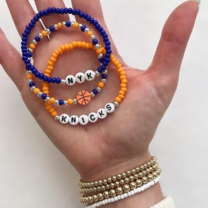 May include: Three beaded bracelets in blue and orange, with silver accents. One bracelet spells "NYK" and another spells "KNICKS" with a basketball charm. Additional gold and white beaded bracelets are also visible.