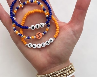 Może przedstawiać: Trzy bransoletki z koralików w kolorze niebieskim i pomarańczowym, ze srebrnymi akcentami. Jedna bransoletka wymawia "NYK", a druga "KNICKS" z zawieszką w kształcie koszykówki. Widoczne są również dodatkowe złote i białe bransoletki z koralików.