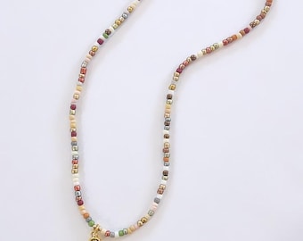 May include: A colorful beaded necklace with a gold-colored pendant in the shape of the letter "S". The necklace features small, square beads in various colors, including white, gold, red, green, and brown. The necklace is displayed on a white background.