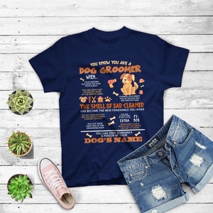 May include: A navy blue t-shirt with a dog groomer design. The text reads "You know you are a dog groomer when..." and lists several humorous signs of being a dog groomer. The design includes a cartoon dog and grooming tools.