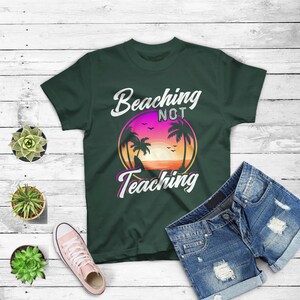 May include: Dark green t-shirt with the text "Beaching Not Teaching" in white and pink. The graphic features a sunset scene with palm trees. Blue denim shorts and pink sneakers are also in the image.