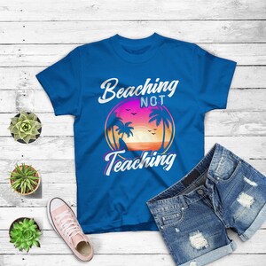 May include: A blue t-shirt with the words "Beaching Not Teaching" in white script. The shirt features a circular graphic of a sunset with palm trees. Denim shorts and a pink sneaker are also in the image.