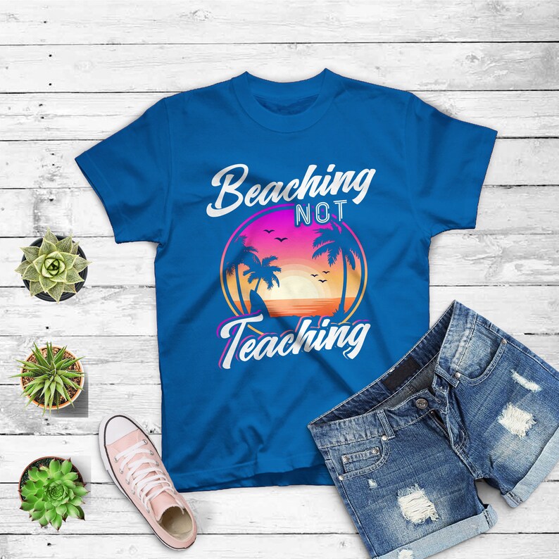 May include: A blue t-shirt with the words "Beaching Not Teaching" in white script. The shirt features a circular graphic of a sunset with palm trees. Denim shorts and a pink sneaker are also in the image.