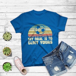 May include: A royal blue t-shirt with a retro sunset graphic featuring a soccer ball and the text "My goal is to deny yours".