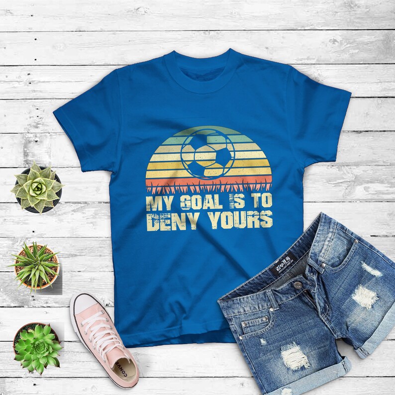 May include: A royal blue t-shirt with a retro sunset graphic featuring a soccer ball and the text "My goal is to deny yours".