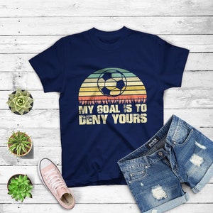 May include: Navy blue t-shirt with a vintage-style graphic of a soccer ball in front of a sunset. The text "My goal is to deny yours" is printed below the graphic.