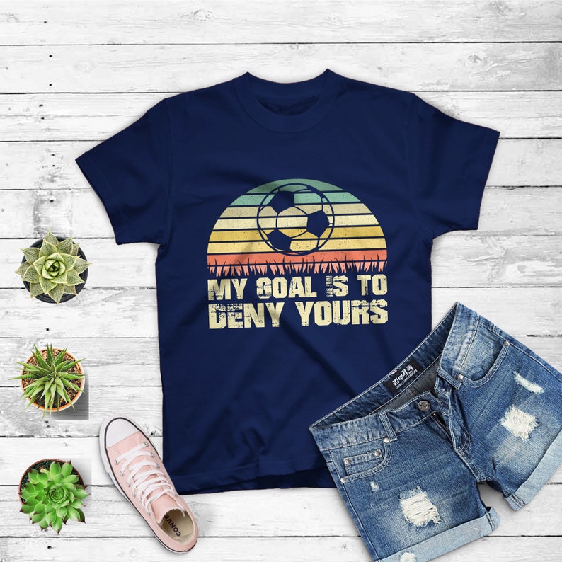 May include: Navy blue t-shirt with a vintage-style graphic of a soccer ball in front of a sunset. The text "My goal is to deny yours" is printed below the graphic.