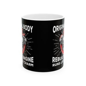 Könnte beinhalten: Schwarze Keramiktasse mit weißem Rand. Die Tasse zeigt eine rote und schwarze Illustration eines Motors mit dem Text "Original Body, Rebuilt Engine Runs Like a Charm".