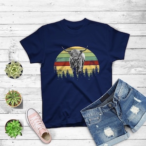 May include: A navy blue t-shirt with a vintage-style graphic of a Highland cow standing in front of a sunset. The graphic is in shades of red, orange, yellow, and green.