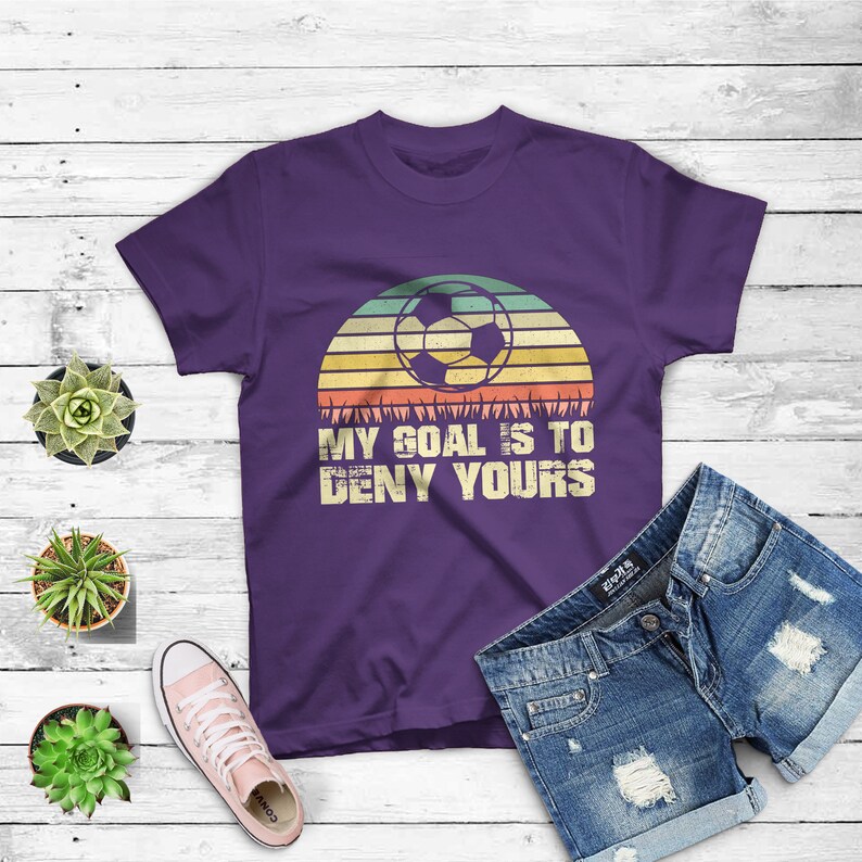 May include: Purple t-shirt with a retro sunset graphic and a soccer ball. The text "My goal is to deny yours" is printed on the shirt.