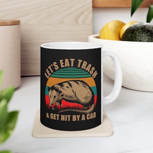 May include: A white ceramic mug with a black design. The design features a retro-style illustration of an opossum with the text "Let's Eat Trash & Get Hit By A Car".
