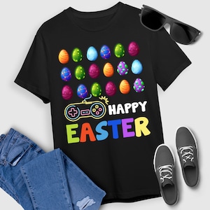May include: Black t-shirt featuring colorful Easter eggs and a video game controller design. The text "HAPPY EASTER" is displayed in multiple colors. Sunglasses, jeans, and sneakers are also visible.