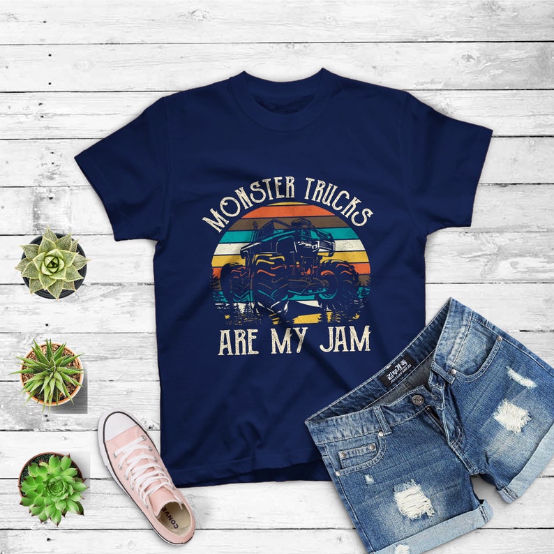 Puede incluir: Una camiseta azul marino con un gr&aacute;fico de estilo vintage de un cami&oacute;n monstruo conduciendo a trav&eacute;s de una puesta de sol. El texto "Monster Trucks Are My Jam" est&aacute; impreso en la camiseta.