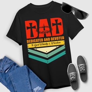 Könnte beinhalten: Schwarzes T-Shirt mit einer roten und weißen Grafik, auf der "DAD DEDICATED AND DEVOTED To God Family & Freedom" steht. Die Grafik zeigt eine Dornenkrone und zwei Kreuze.