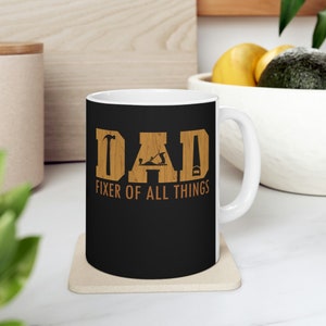 May include: White ceramic mug with a black background and brown wood-grain design. The mug features the word "DAD" with a hammer, saw, and plane graphic. The text "Fixer of All Things" is below the word "DAD".