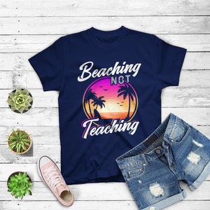 May include: Navy blue t-shirt with the text "Beaching Not Teaching" in white and pink script. The design features a sunset scene with palm trees. A pair of denim shorts and a pink sneaker are also visible.