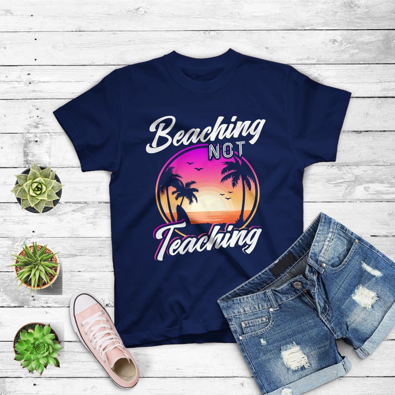 May include: Navy blue t-shirt with the text "Beaching Not Teaching" in white and pink script. The design features a sunset scene with palm trees. A pair of denim shorts and a pink sneaker are also visible.