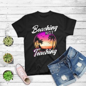 May include: Black t-shirt with the text "Beaching Not Teaching" in white and pink script. The shirt features a sunset graphic with palm trees. The shirt is paired with denim shorts and a pair of pink sneakers.
