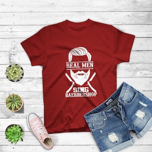 May include: Red t-shirt with a white graphic of a man's face with a beard and the text "REAL MEN SING BAERBERSHOP".