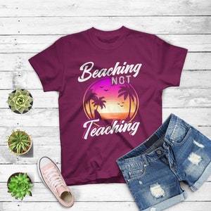 May include: A maroon t-shirt with the words "Beaching Not Teaching" in white script. The graphic features a sunset scene with palm trees. Blue denim shorts and a pair of pink sneakers are also in the image.