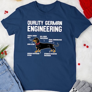 May include: A navy blue t-shirt with a white dachshund illustration and the text "Quality German Engineering". The dachshund is labeled with humorous anatomical features, such as "frontal sensor units", "50% larger than average", "visual communication device", "audio alarm", "lowered suspension system", and "optimized excavation mechanism".