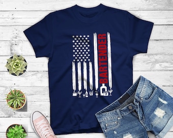 May include: A navy blue t-shirt featuring a white and red American flag design. The text "BARTENDER" is printed vertically on the right side of the flag. The flag is stylized with white brushstrokes. There are small icons of drinks below the flag.
